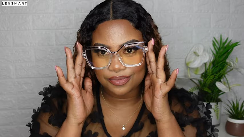 Mimi Manaso models the Marly glasses by Lensmart, adjusting the large, clear square frames on her face. The glasses have a modern, classy look, and she smiles at the camera, showcasing their comfortable and playful style.